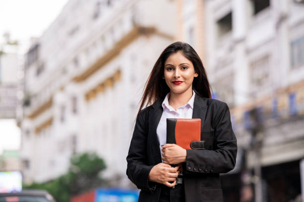 Outdoor Portrait of young Indian student outside campus building, she is holding notebook or diary and looking at camera.
