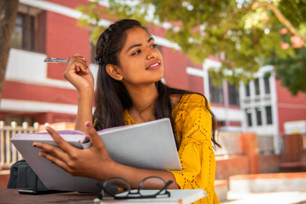 Portrait of beautiful Asia, Indian female college student reading a book and doing preparation for the exam at campus. She is holding a book and contemplating while looking away with a smile.